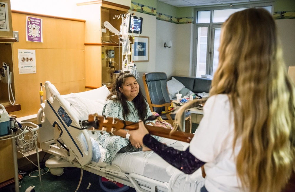 A volunteer musician plays for a paitent at their bedside. She is smiling as the music is being played