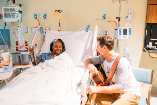 Charles Esten laughs with an adult Black patient who is in her hospital bed. Charles has his guitar to the side