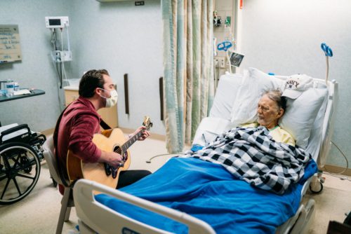 Alex Hall is wearing a face mask and sitting bedside to a hospitalized veteran. He is playing his guitar as the Vet listens