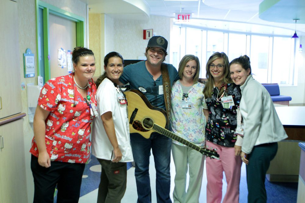 Caregivers and hospital workers pose with Jerrod Neimann, guitar in hand, at a MOC Bedside program