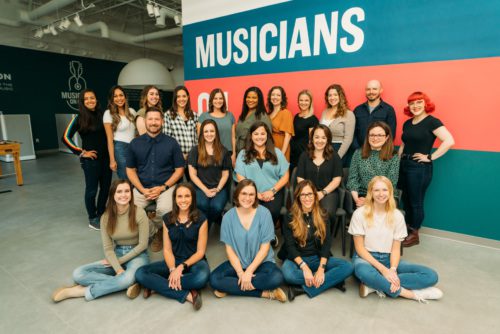 musicians on call staff members are positioned in three rows, all with welcoming smiles. The front row is seated on the floor, the middle is seated in chairs, and the third is standing behind