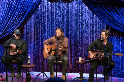 The three performers, Dierks Betley, Jon Nite and Charles Esten, sit on stools and have acoustic guitars in their hands. They are playing together at Concert For Caregivers, a show celebrating hospital workers