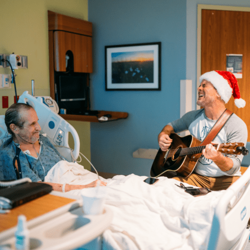 Charles Esten, wearing a santa hat, is playing guitar bedside for a hospitalized patient. The patient is smiling, showing gratitude for this moment of music and joy